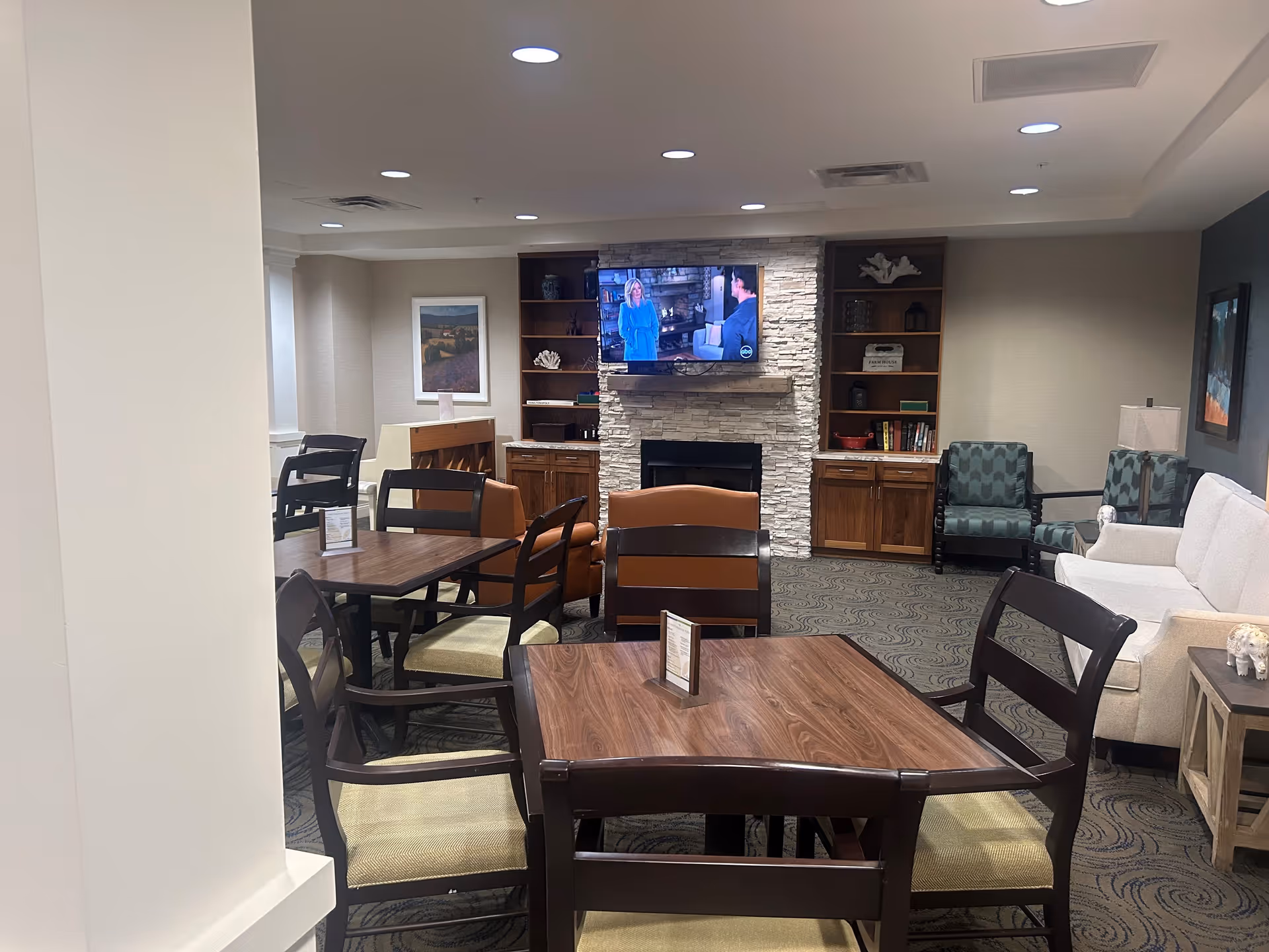 A senior living facility common area with multiple wooden tables and chairs arranged for seating. In the background, there is a stone fireplace with a mounted flat-screen TV showing a scene from a TV show. Surrounding the fireplace are built-in wooden shelves and cabinets with decorative items. The room also features a white couch, patterned armchairs, a side table with a lamp, and framed artwork on the walls. The ceiling has recessed lighting.