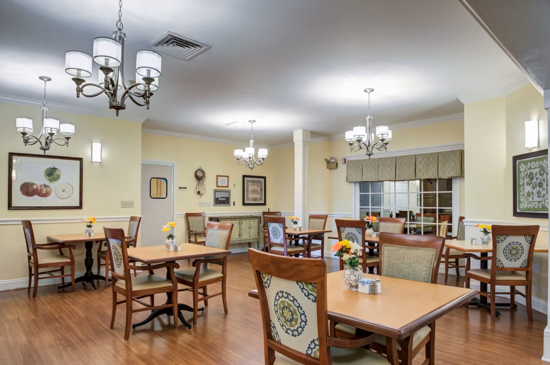 Bright dining room with wooden tables and upholstered chairs, chandeliers, and floral centerpieces.