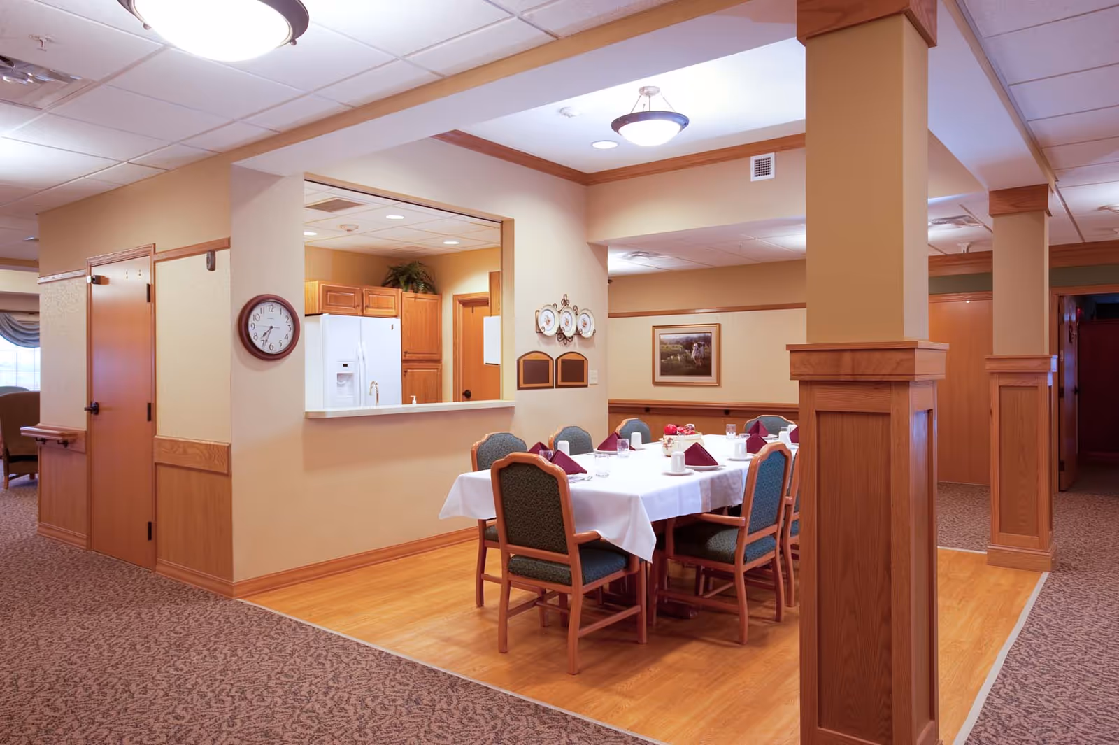 A dining area in a senior living facility with a rectangular table covered with a white tablecloth, set with cups, glasses, and folded burgundy napkins. The room has wooden pillars, beige walls, and a clock on the wall. A kitchen area with wooden cabinets and a white refrigerator is visible through a pass-through window.