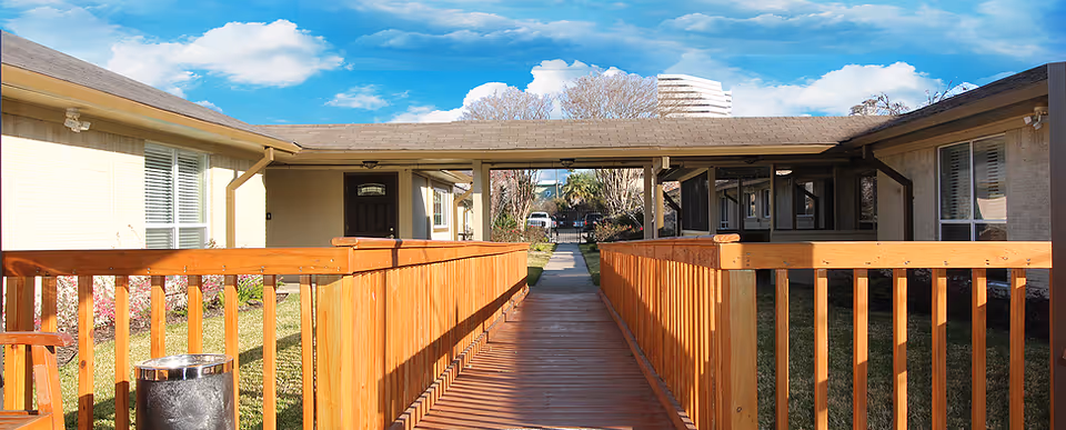 Outdoor view of a senior living facility showing a wooden ramp with railings leading to the entrance of a building. The building has beige walls and multiple windows with white blinds. The sky is blue with some clouds, and there is greenery on both sides of the ramp.