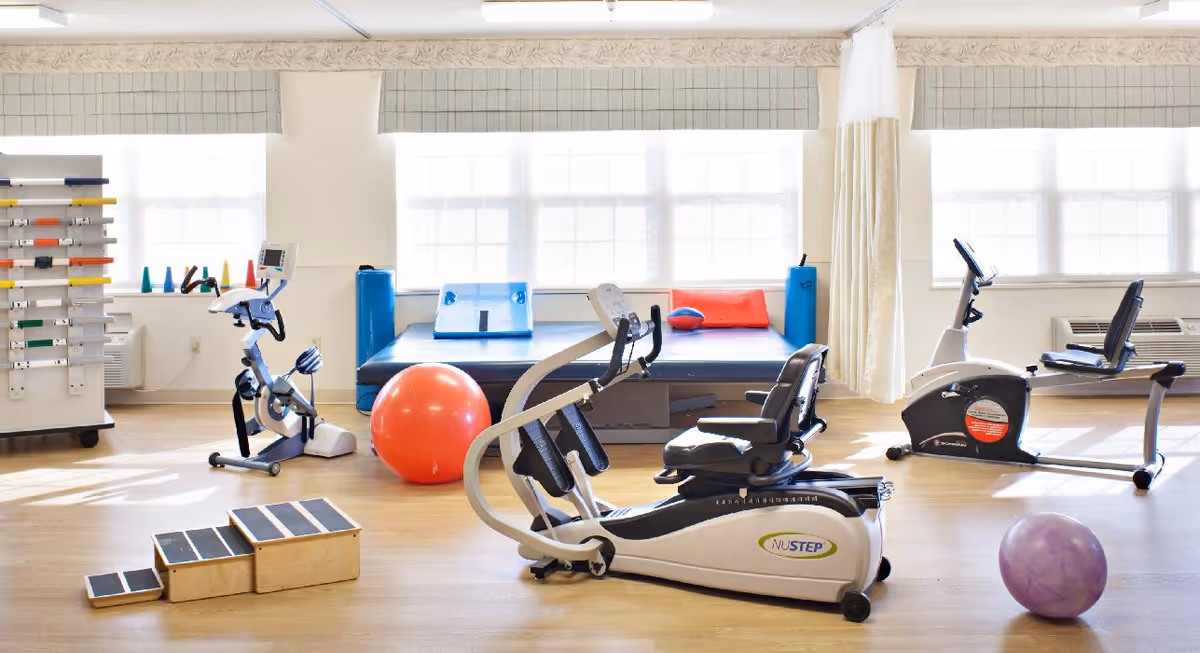 A bright and spacious physical therapy or exercise room with various fitness equipment including stationary bikes, a large exercise ball, a purple smaller ball, a padded exercise platform with cushions, and wooden step platforms. The room has large windows with blinds allowing natural light to fill the space.