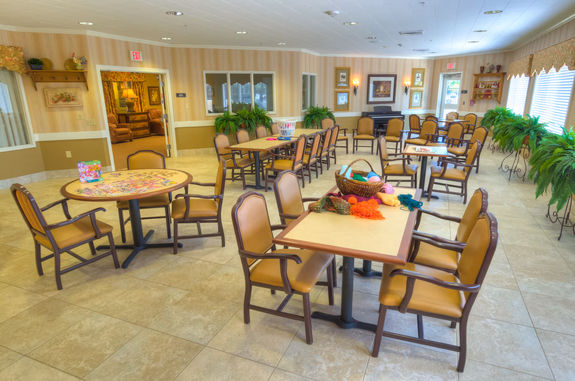 Bright communal activity/dining room with multiple tables and chairs, potted plants, and craft items on the tables.