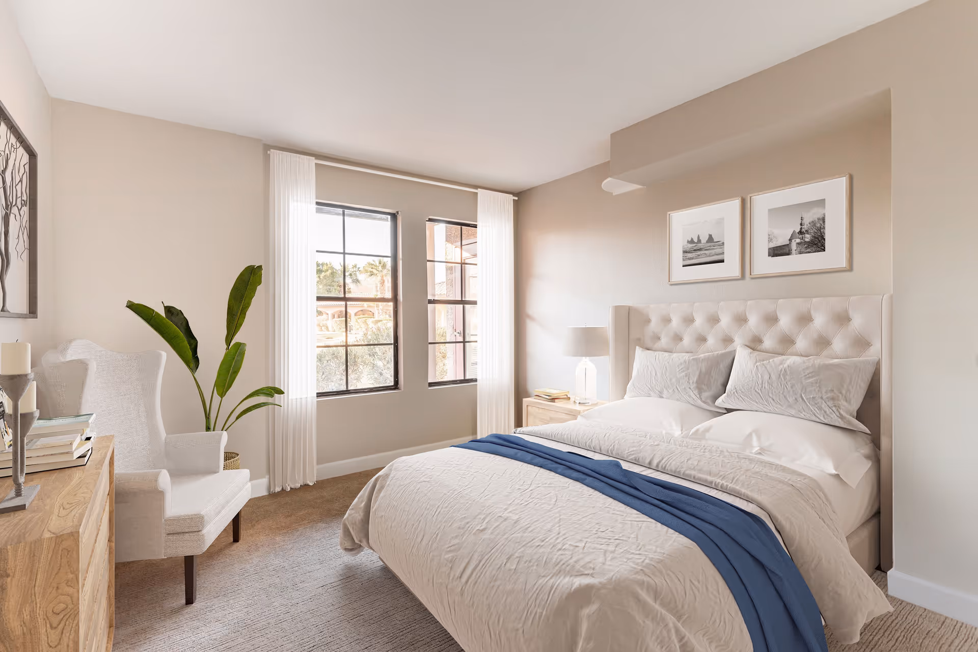 A bright and cozy bedroom with a large bed featuring a tufted headboard, white bedding, and a blue throw blanket. There are two framed black and white pictures on the wall above the bed. A wooden nightstand with a lamp and books is beside the bed. A white armchair and a wooden dresser with books and a candle are on the left side of the room. Two windows with white curtains allow natural light to fill the space. A green potted plant is placed near the windows.