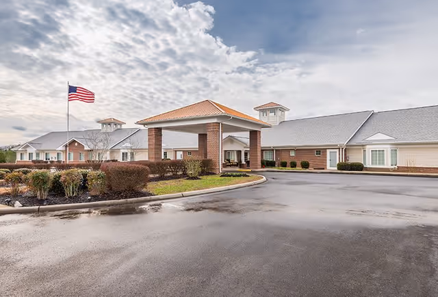 Front entrance of a single-story senior living facility with a covered porte-cochere, American flag, and landscaped driveway under a cloudy sky.
