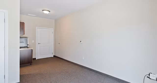 Empty room with beige walls and brown carpet flooring, a closed white door, and a small kitchenette area with dark cabinets and a microwave on the left side.