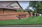 Exterior view of Brewster Place facility showing a brick wall with the Brewster Health & Wellness Services sign, a manicured lawn, and part of the building with a sloped roof in the background.