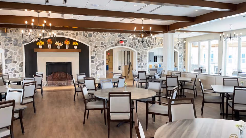 Bright communal dining room with round and square tables, chairs, chandeliers and a stone fireplace.