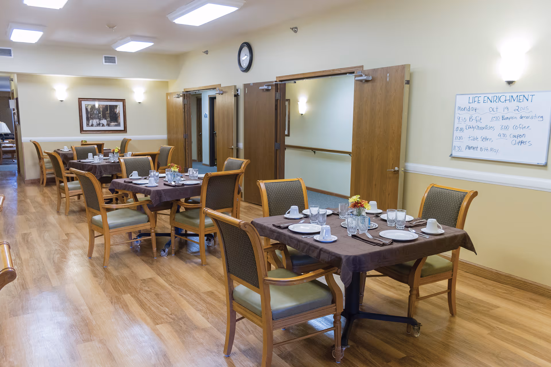 A dining room in a senior living facility with several tables covered with brown tablecloths, each set with plates, cups, glasses, and silverware. There are wooden chairs with green cushions around the tables. The room has light yellow walls, wooden flooring, and a clock above two open double doors. A whiteboard on the wall lists life enrichment activities for Monday, October 19, 2015.