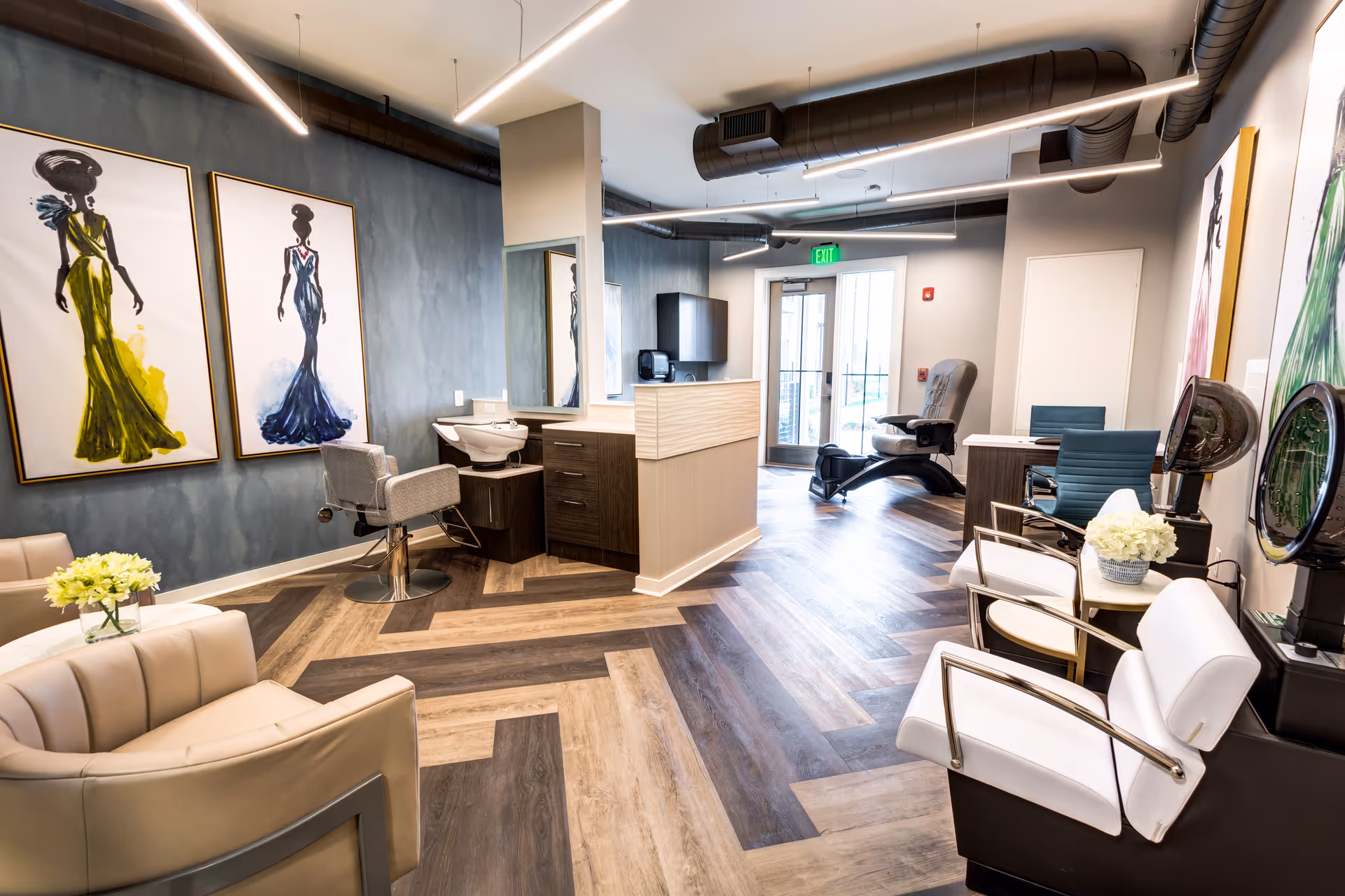 Interior of a modern salon area in a senior living facility with styling chairs, hair washing station, hair dryers, and comfortable seating. The room features stylish wooden flooring, contemporary lighting, and large fashion artwork on the walls.