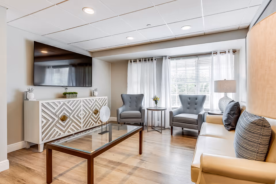 A bright and modern living room with a large flat-screen TV mounted on the wall above a decorative white cabinet. The room features two gray armchairs with button tufting, a beige sofa with striped and solid pillows, a glass coffee table with a wooden frame, and a side table with a lamp and a small flower arrangement. White curtains frame a large window letting in natural light, and the floor is a light wood laminate.
