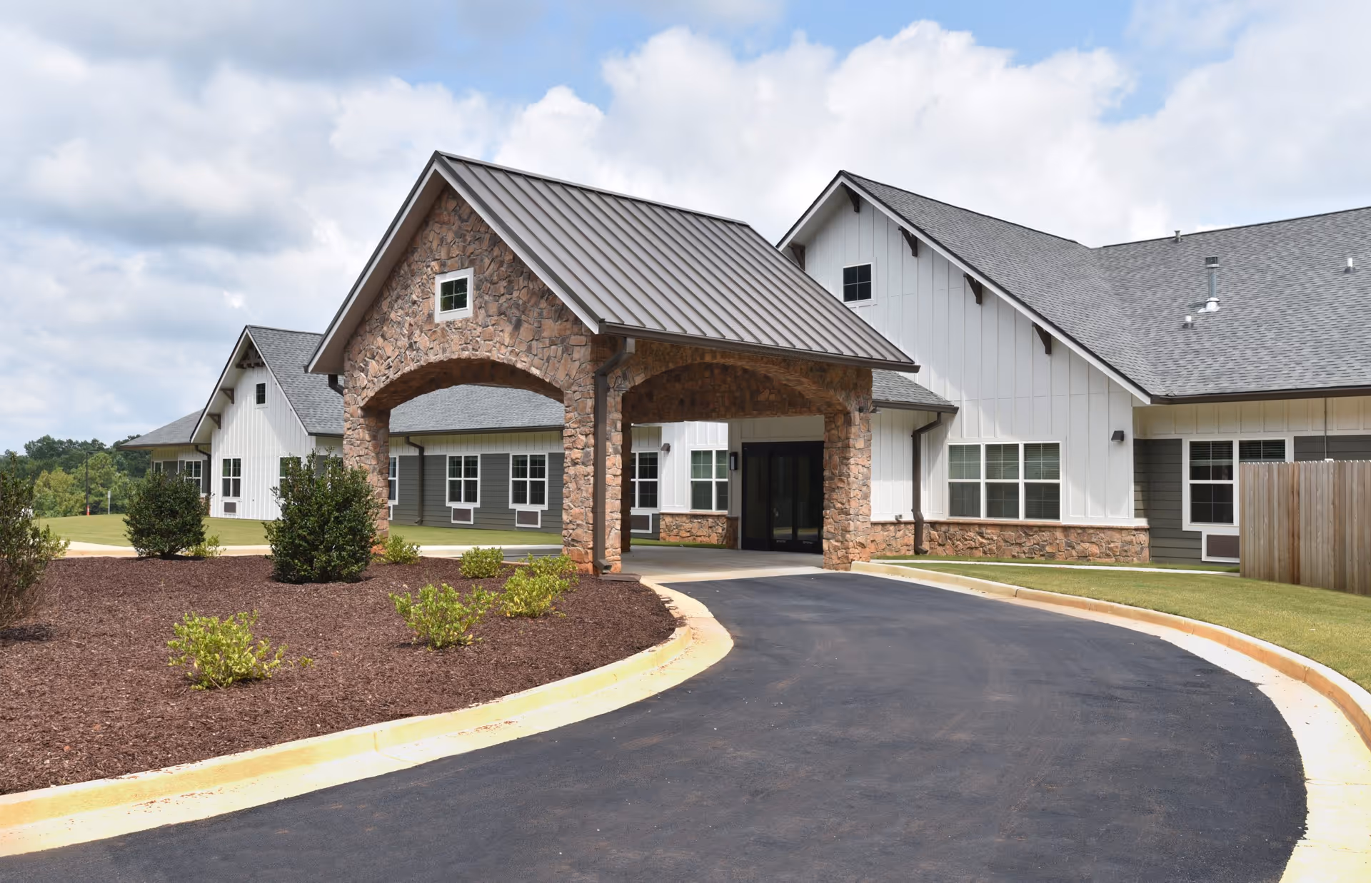 Exterior view of a senior living facility building with a covered entrance made of stone and a paved driveway leading up to it. The building has white siding with stone accents and multiple windows under a partly cloudy sky.