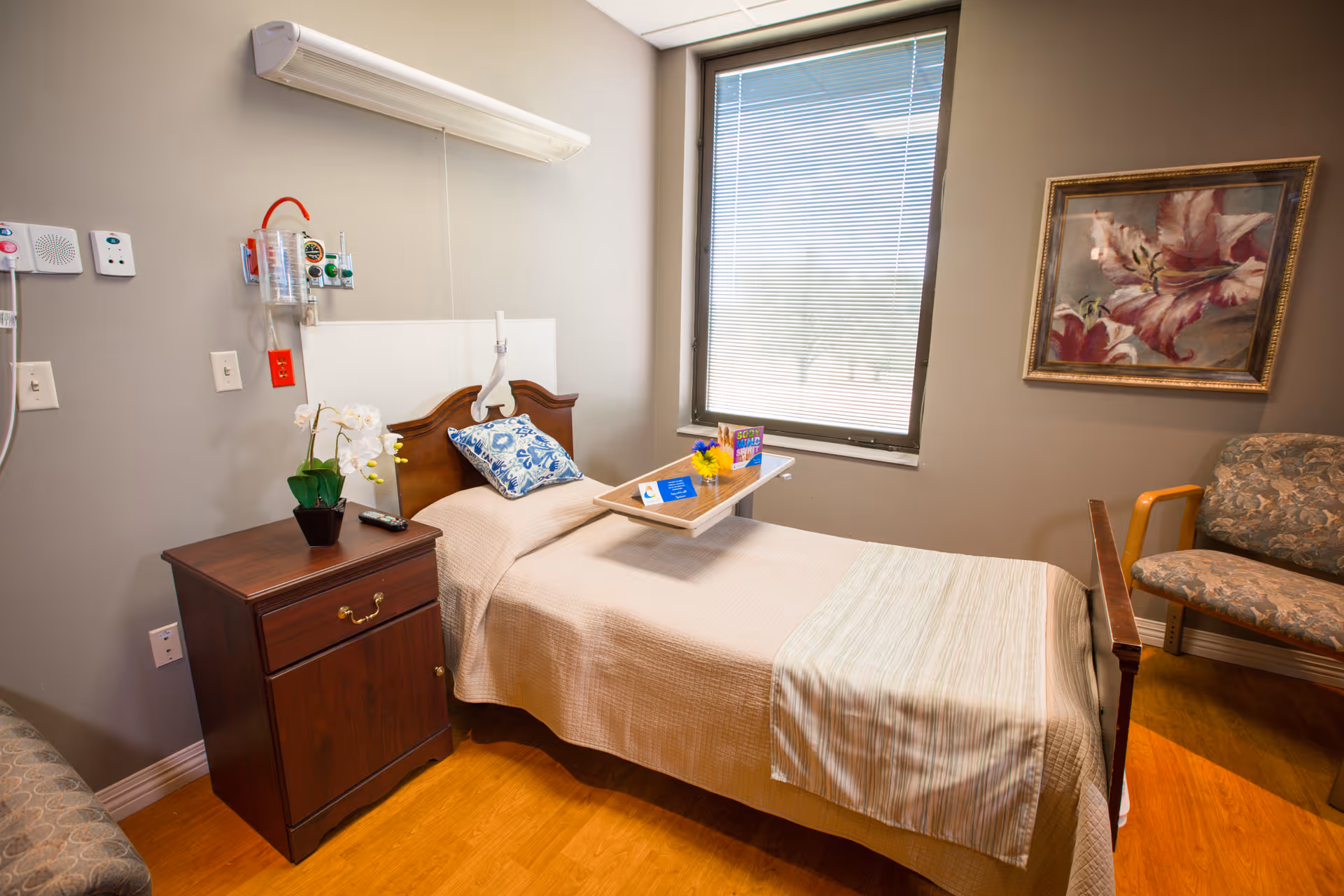 A single bed in a senior care facility room with a beige bedspread and a blue patterned pillow. A wooden nightstand with a potted white orchid and a remote control is beside the bed. A tray with a small bouquet of flowers and a card is placed on the bed. The room has a window with blinds, a floral painting on the wall, and a cushioned chair with wooden armrests.