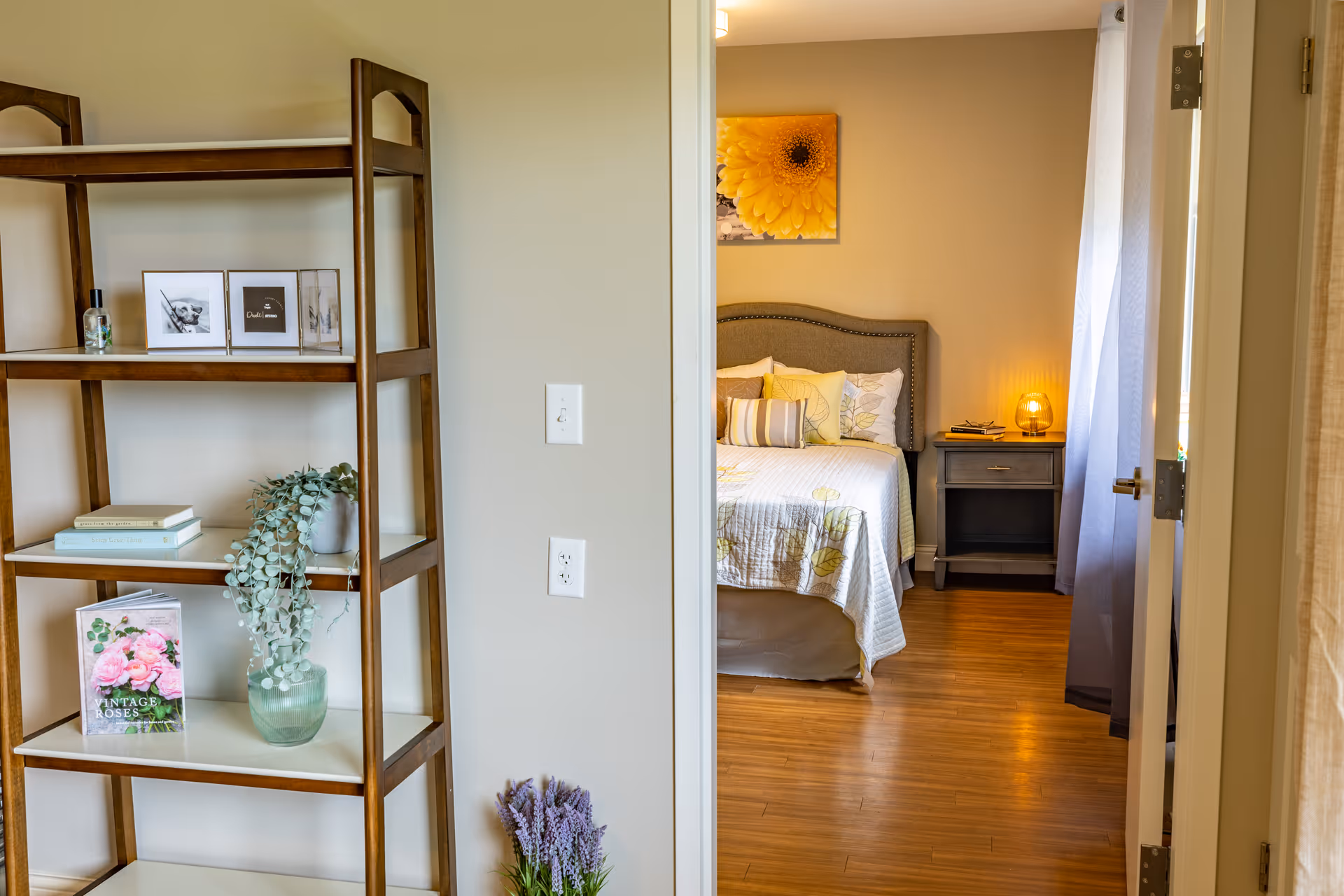 Open doorway showing a decorated bedroom with a bed, nightstand and lamp, and a wooden shelving unit with plants and books in the foreground.