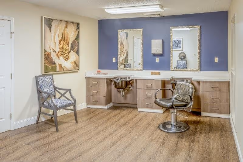 Interior room with a salon-style sink and chair, two mirrors mounted on a blue accent wall above a countertop with cabinets, a patterned chair against a beige wall, and a large floral painting.