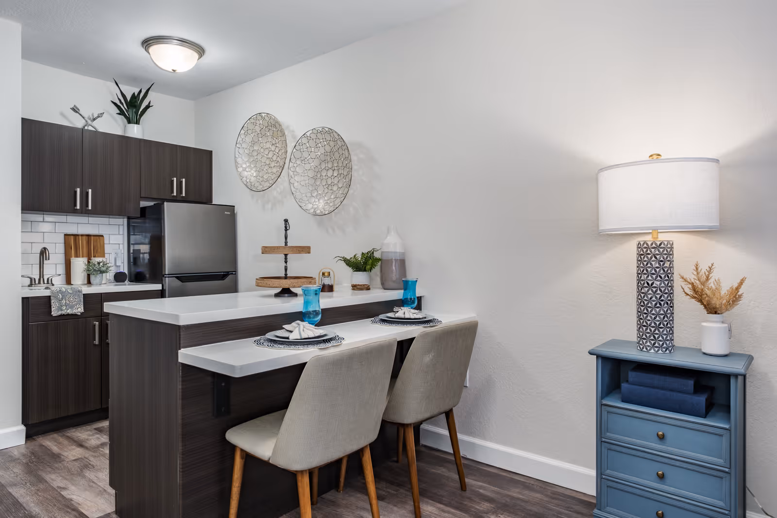 Modern kitchen area with dark wood cabinets, a stainless steel refrigerator, a white countertop with two place settings including blue glasses, and two beige chairs. On the right side, there is a blue side table with a decorative lamp and a small vase with dried plants. Two round decorative wall hangings are mounted on the white wall above the countertop.