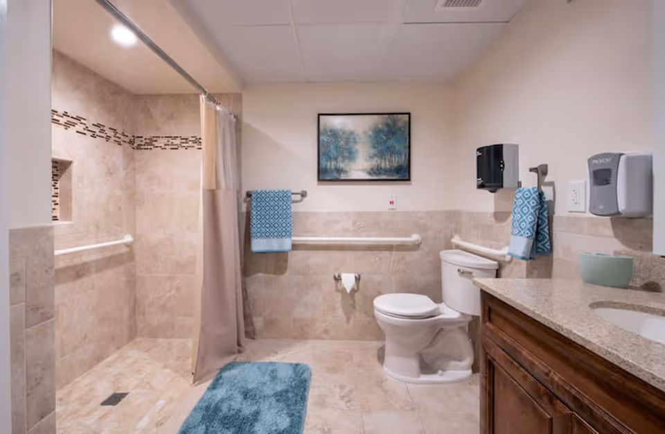 A clean and accessible bathroom with beige tiled walls and floor, featuring a walk-in shower with a curtain, a white toilet, a wooden vanity with a granite countertop, blue patterned towels hanging on grab bars, a blue bath mat on the floor, a wall-mounted soap dispenser, and a framed painting of a forest scene on the wall.