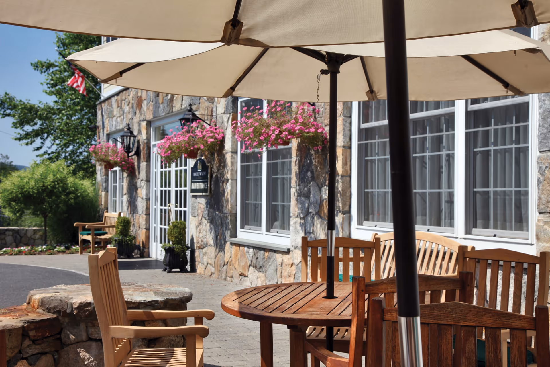 Outdoor seating area with wooden chairs and a round table under large beige umbrellas in front of a stone building with windows and hanging flower baskets. An American flag is visible near the entrance door.