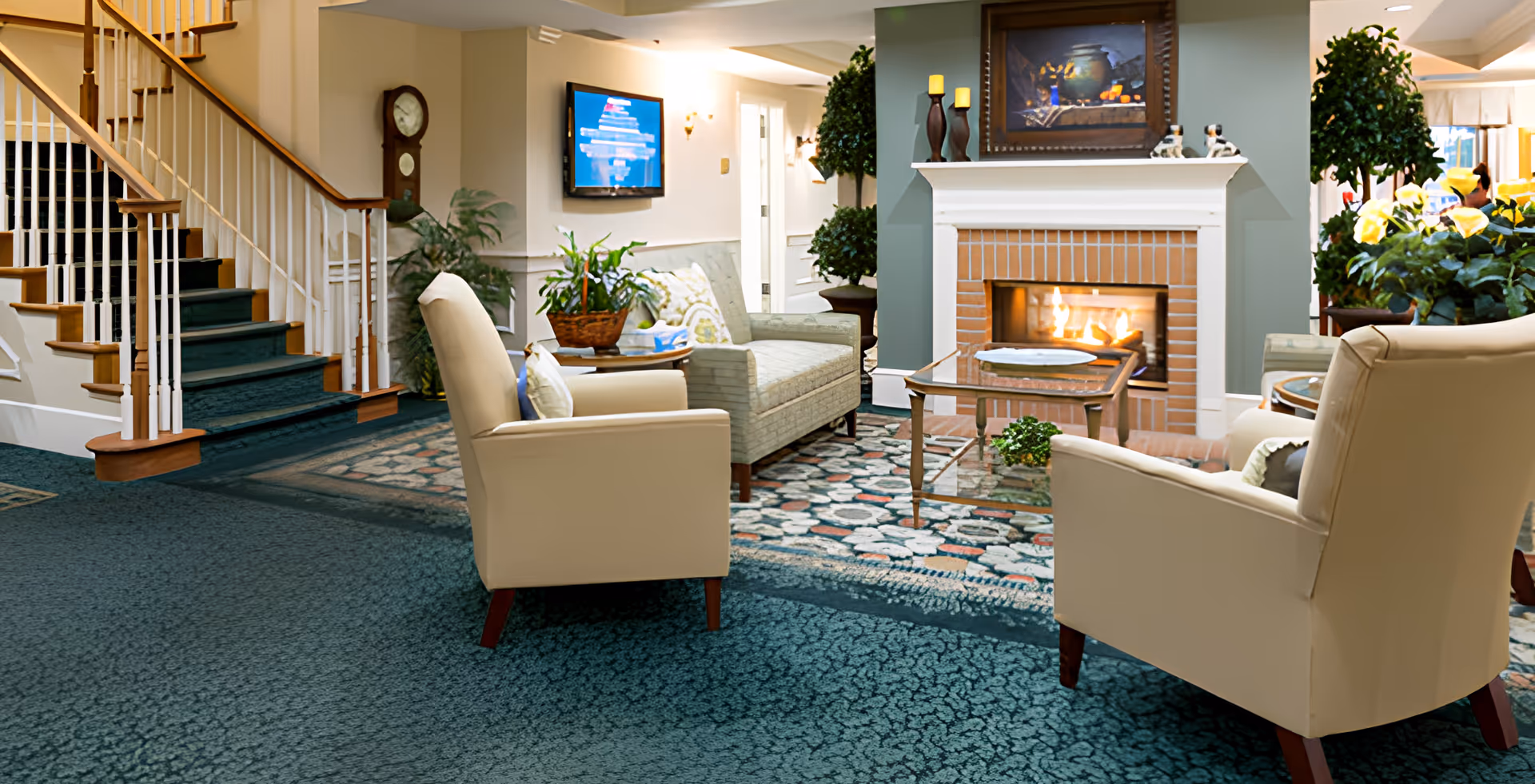 Cozy lobby area with armchairs and a sofa arranged around a lit fireplace, a coffee table, plants, and a nearby staircase.