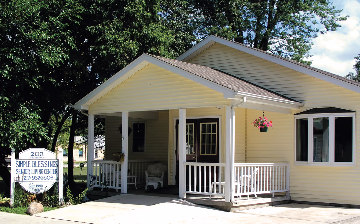 Exterior view of Simple Blessings Senior Living Center, a single-story yellow building with a covered porch, white railings, and outdoor chairs. A hanging flower pot with pink flowers is visible on the porch. There is a sign in front of the building displaying the facility name, phone number, and establishment year. Trees and a partly cloudy sky are in the background.