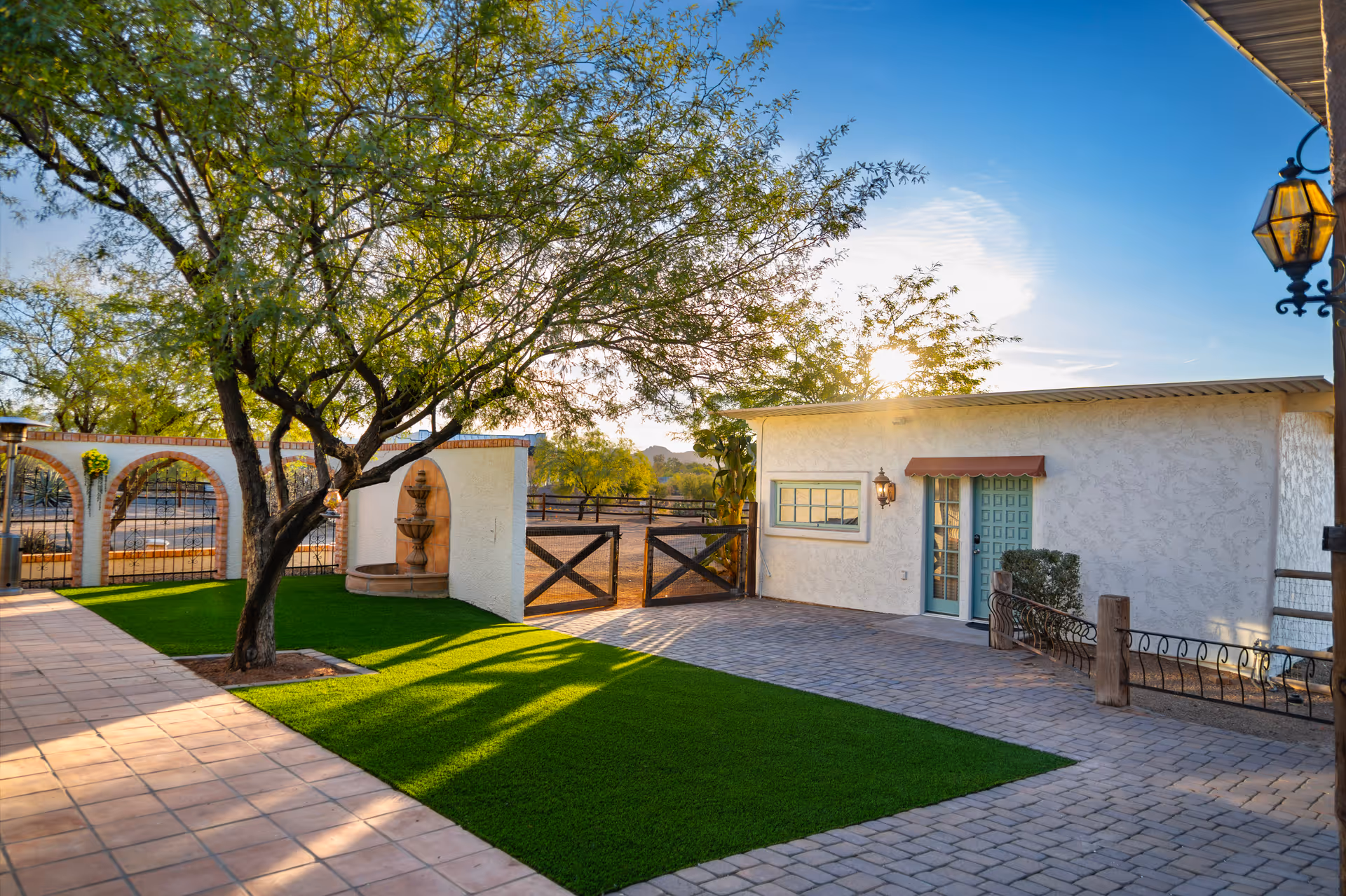 Outdoor courtyard area with a tree casting shadows on green grass and tiled ground. There is a white building with a teal door and window, a decorative wall fountain, and a wooden gate leading to an open area with trees and mountains in the background under a blue sky with the sun shining.