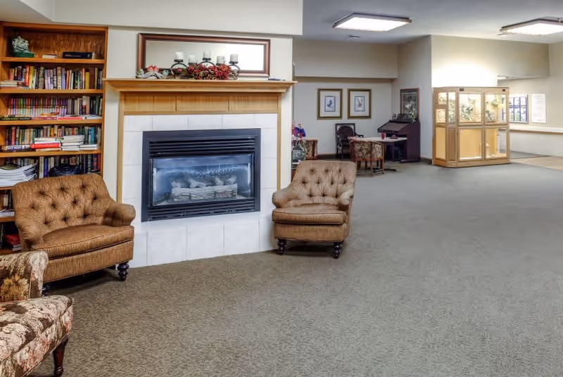 A cozy living room area in a senior living facility featuring a fireplace with a wooden mantel decorated with candles and flowers. Two brown upholstered armchairs are placed on either side of the fireplace. To the left, there is a built-in wooden bookshelf filled with books. In the background, there are additional seating areas with chairs and framed artwork on the walls, along with a large display cabinet and windows letting in natural light.