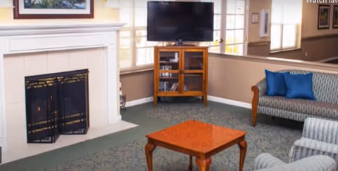 A cozy living room area with a fireplace on the left, a flat-screen TV on a wooden stand in the center, a patterned sofa with blue cushions on the right, and a wooden coffee table in the middle. The room has large windows with blinds and framed pictures on the walls.