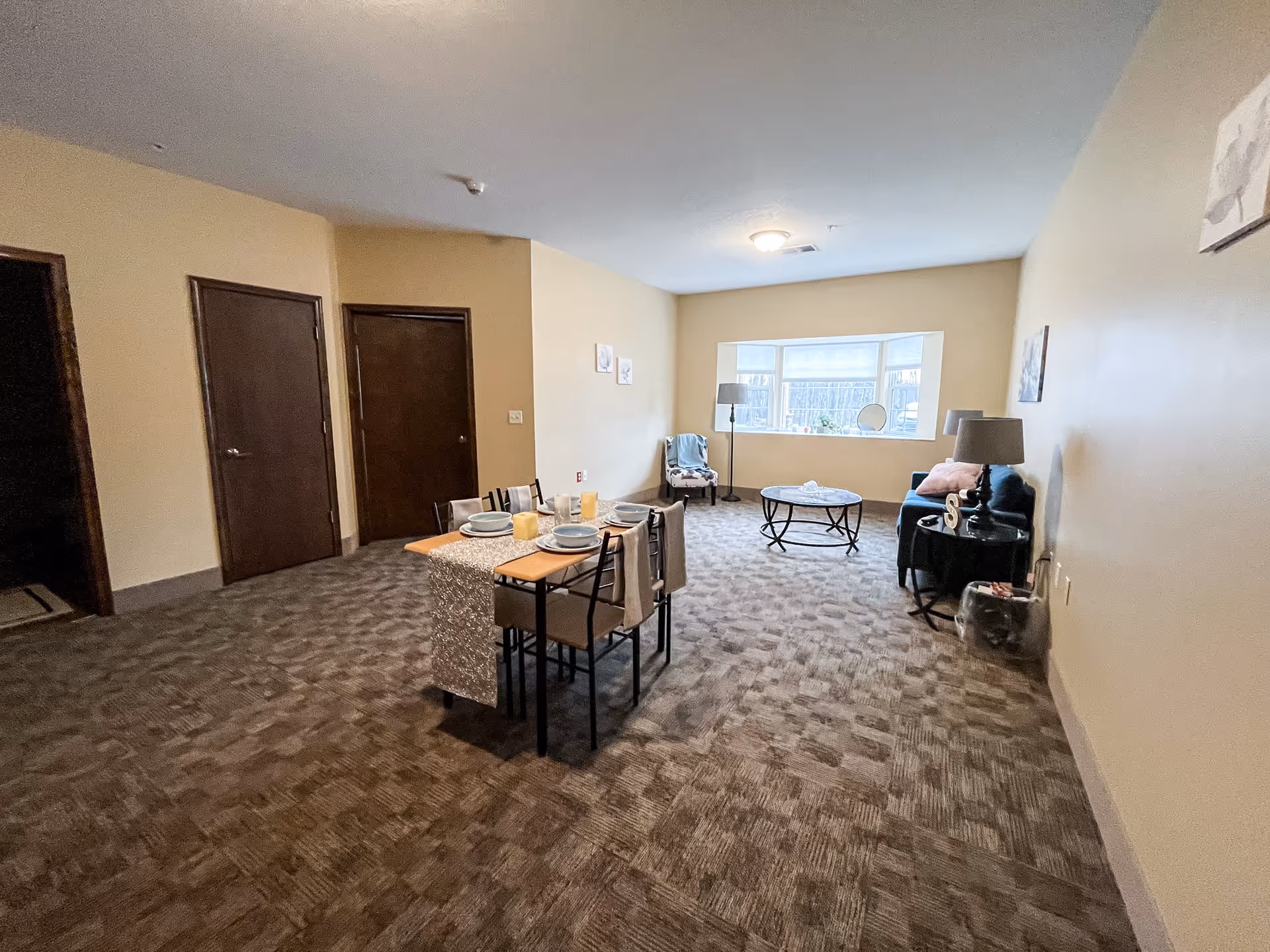 A spacious room with beige walls and carpeted floor featuring a small dining table set for four with plates and candles, a sitting area with a blue couch, a round coffee table, a chair with a blanket, a floor lamp, and a window letting in natural light. Two closed wooden doors are visible on the left side of the room.