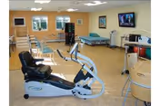 A rehabilitation room with exercise equipment including a recumbent bike, parallel bars for walking practice, a hospital bed, and various physical therapy tools. The room has beige walls, a window, and a television mounted on the wall.