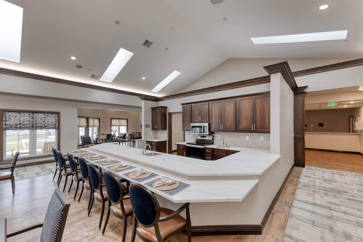 A spacious senior living facility kitchen with a large white marble countertop island surrounded by chairs set with placemats and napkins. The kitchen features dark wood cabinets, stainless steel appliances, and a white subway tile backsplash. Skylights and recessed lighting illuminate the room, with a dining area visible in the background through large windows.