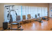 Small fitness room with a multi-station strength machine, stationary bike and rowing machine, and a row of chairs along a large window.