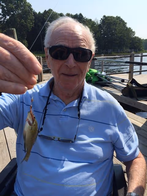 An elderly man wearing sunglasses and a light blue polo shirt is sitting on a wooden dock by the water, holding up a small fish he caught. Trees and a kayak are visible in the background.