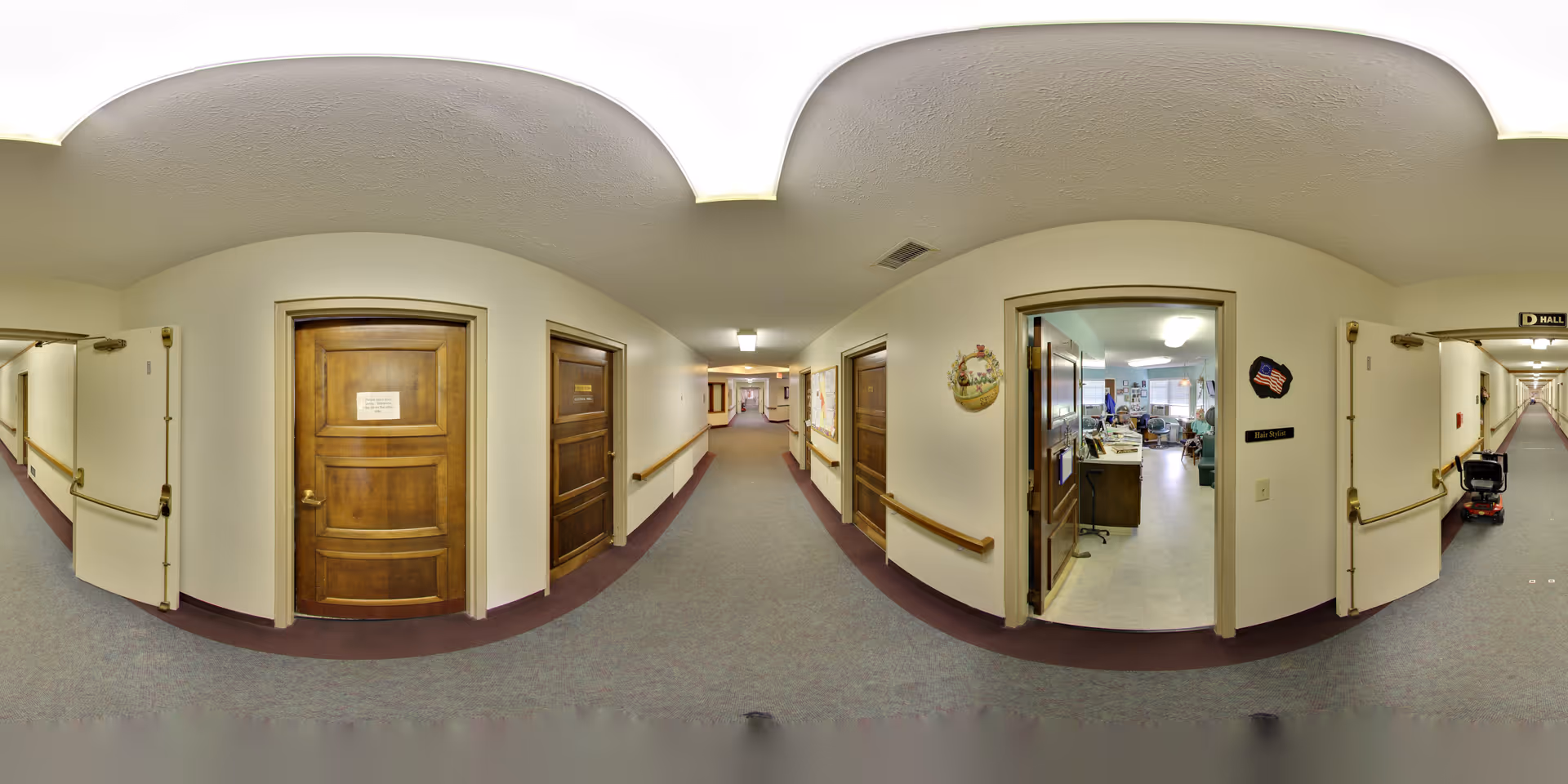 A wide-angle view of a hallway in a senior living facility with beige walls, wooden doors, handrails along the walls, and carpeted floors. One door is open, revealing an office space with desks, chairs, and people working inside. The hallway extends into the distance with more doors and lighting fixtures on the ceiling.