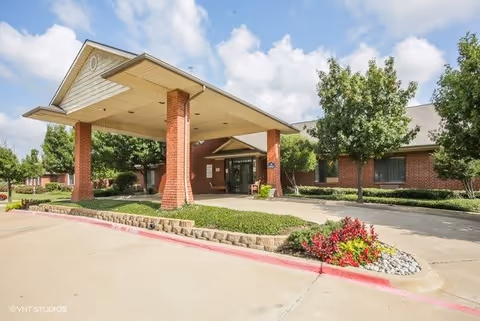 Front entrance of a brick senior living building with a covered porte-cochere, landscaping, and driveway under a blue sky.