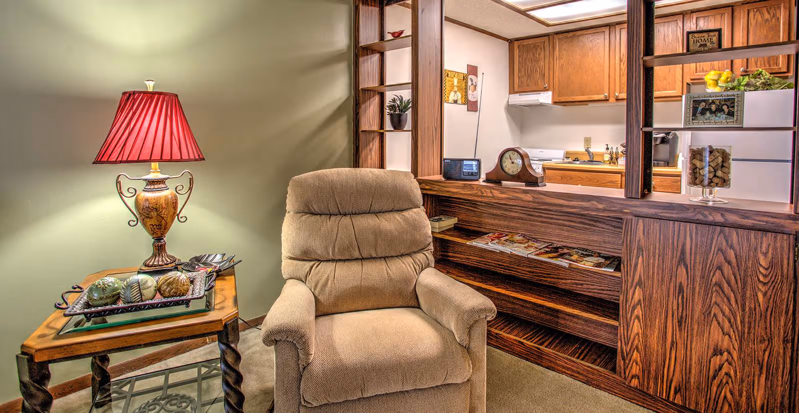 A cozy living area with a beige recliner chair next to a wooden side table holding a decorative lamp with a red shade and a tray with decorative balls. Behind the chair is a wooden partition with shelves containing magazines, a clock, and a radio. The partition separates the living area from a kitchen with wooden cabinets, a white stove, and a refrigerator.