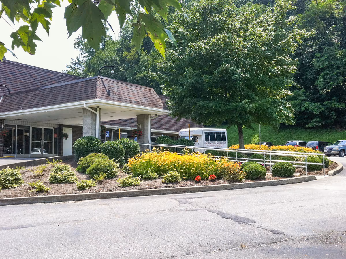 Exterior view of Charleston Healthcare Center showing the entrance with a covered drop-off area, surrounded by landscaped bushes and yellow flowers. A white shuttle van is parked near the entrance, and several cars are visible in the background. Trees provide shade around the building.