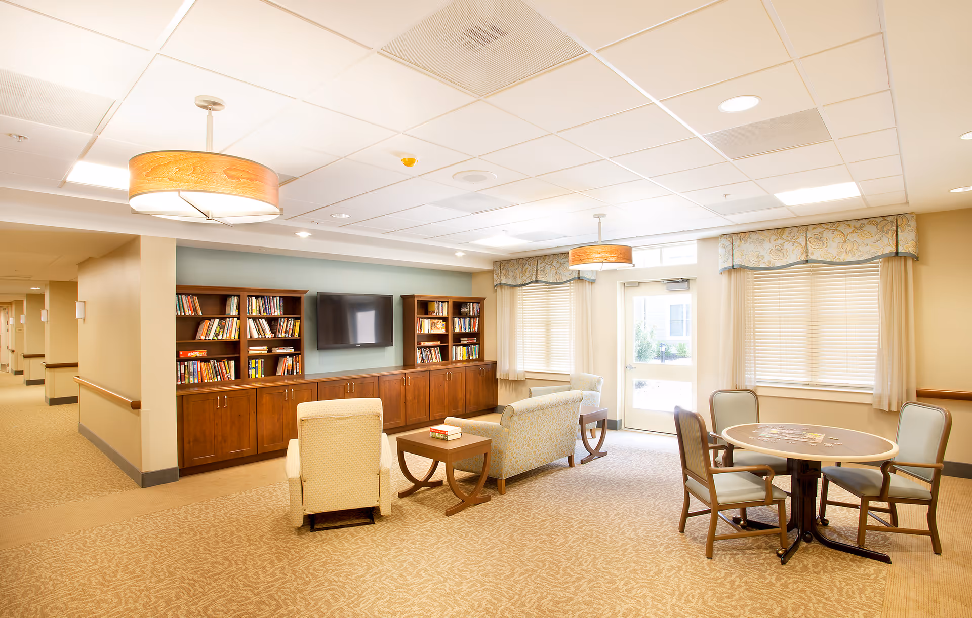 Bright communal living room with bookshelves and TV, several armchairs and a coffee table, and a round game table by the windows.