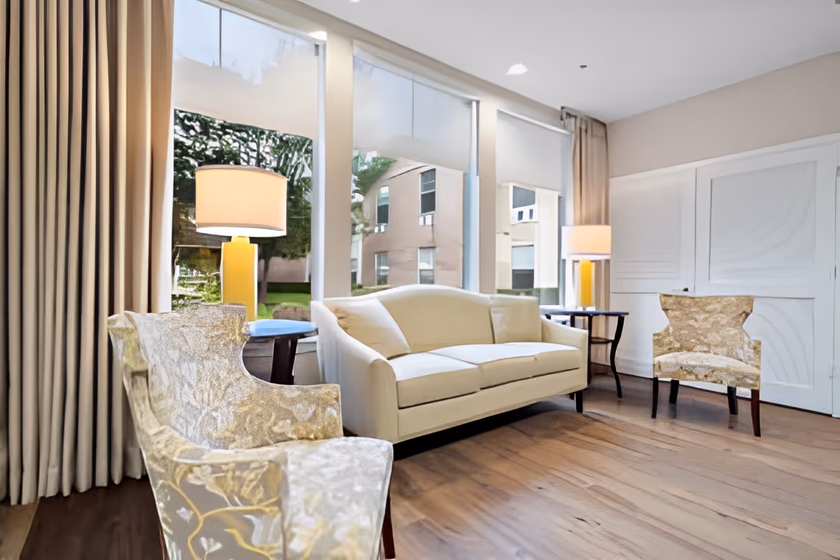 Sunlit lounge with a beige sofa, two patterned armchairs, side tables and lamps in front of large windows.
