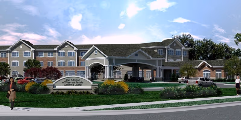 Front exterior of a multi-story assisted living building with a covered entrance, landscaping, sign, and parked cars.