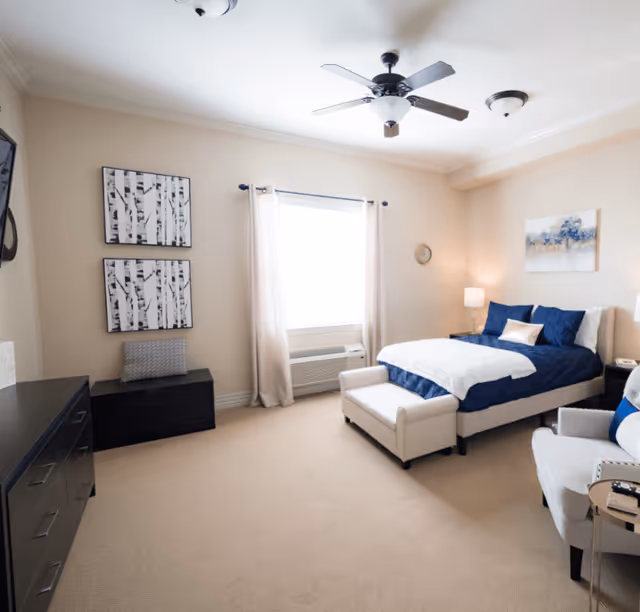A bright and spacious bedroom with beige walls and carpet. The room features a bed with navy blue bedding and white accents, a white bench at the foot of the bed, a white armchair with a blue pillow, a black dresser, and two framed black and white birch tree prints on the wall. A window with light curtains lets in natural light, and a ceiling fan is mounted above.