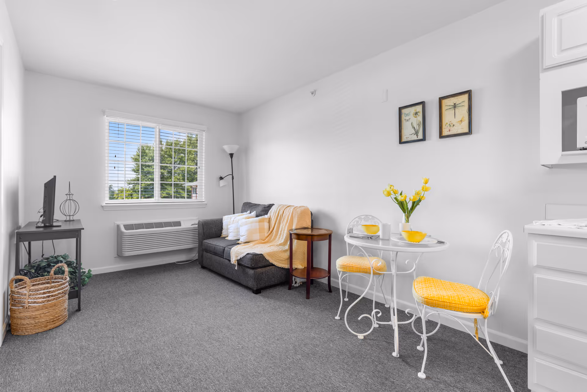 A bright and cozy living area in an assisted living facility featuring a gray loveseat with white and yellow pillows and a yellow throw blanket. Next to the loveseat is a small round wooden side table and a tall floor lamp. A small white metal dining table with two matching chairs with yellow cushions is set with yellow bowls and a vase of yellow tulips. The room has a large window with white blinds, a wall-mounted air conditioning unit, and white walls decorated with two framed botanical prints. A small TV sits on a black metal stand with a decorative item and a woven basket with greenery underneath.