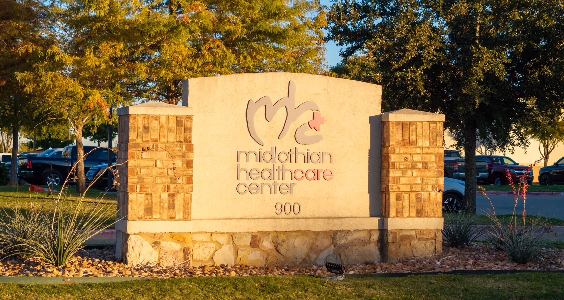 Stone and brick sign for Midlothian Healthcare Center with the address number 900, surrounded by landscaping and trees with parked cars in the background.