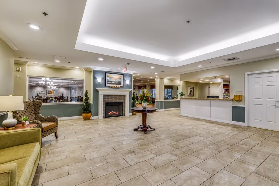 Spacious and well-lit senior living facility common area with tiled floor, a fireplace with a framed picture above it, a round wooden table with a flower vase in the center, a green sofa and a patterned armchair with a side table and lamp, a reception desk on the right, and a large window showing a dining area with tables and chairs.