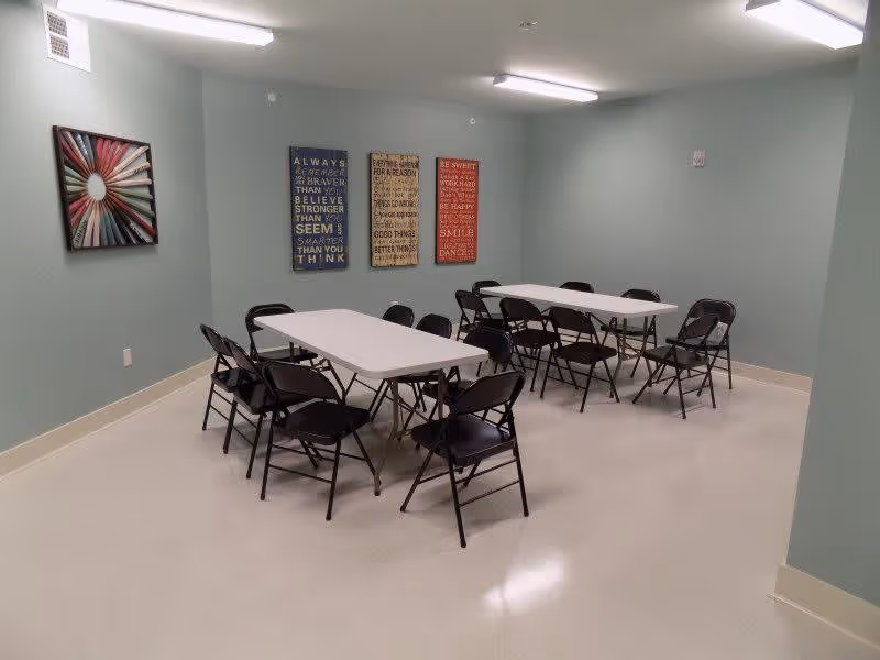 A simple room with light green walls and white floor, containing two rectangular white tables surrounded by black folding chairs. Three framed inspirational quote posters and one colorful abstract artwork hang on the walls. The room is lit by fluorescent ceiling lights.