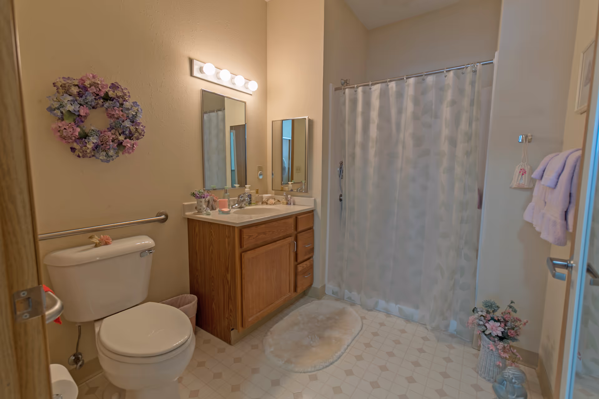 A bathroom with a toilet, wooden vanity with a sink and mirror, a shower with a patterned curtain, a floral wreath on the wall, and decorative flowers on the floor. Towels hang on a rack near the door.