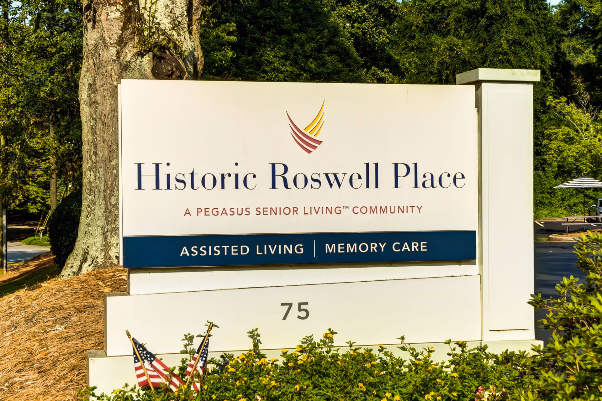 Outdoor sign for Historic Roswell Place, a Pegasus Senior Living community offering assisted living and memory care, surrounded by greenery and small American flags.