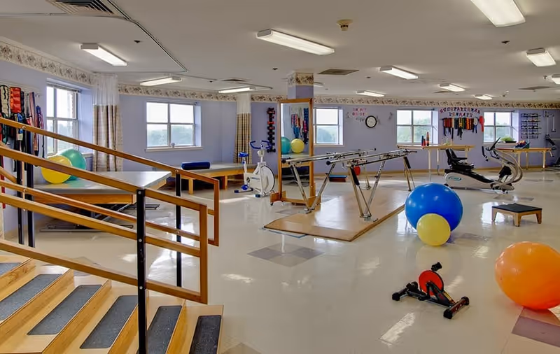 A spacious rehabilitation room with exercise equipment including parallel bars, stationary bikes, large exercise balls, and various therapy tools. The room has multiple windows letting in natural light, light purple walls, and a wooden handrail along a small staircase.