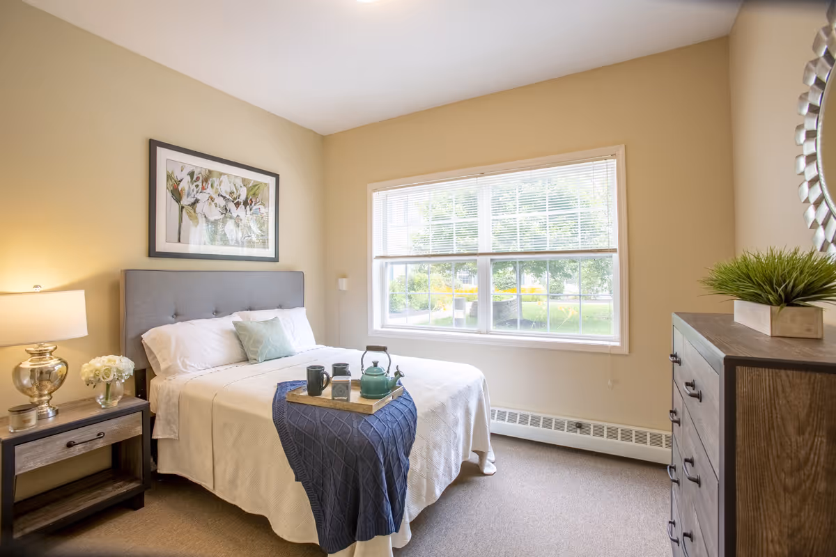 A bright and cozy bedroom with a large window letting in natural light. The room features a neatly made bed with white bedding, a blue throw blanket, and a tray with a teapot and cups. There is a nightstand with a lamp and flowers on the left side of the bed, a framed floral artwork above the headboard, and a wooden dresser with a decorative plant and a round mirror on the right side.