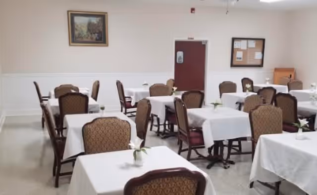 A dining room with multiple tables covered in white tablecloths, each table having a small flower vase with a white flower. The room has several upholstered chairs with patterned backs, a painting on the wall, a bulletin board, and a closed door.