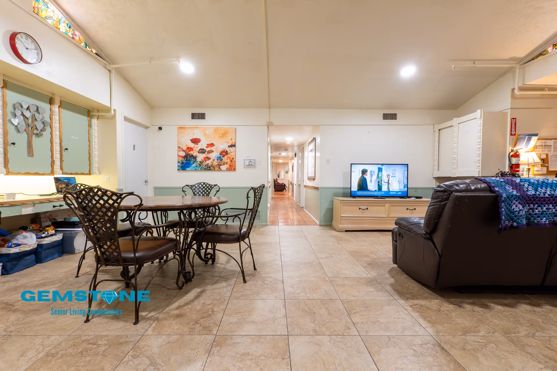 Interior view of a senior living facility common area with a tiled floor. On the left, there is a round table with four metal chairs. Behind the table, there are cabinets with decorative artwork and a clock on the wall. In the center background, a hallway extends further into the building. On the right side, there is a television on a wooden stand showing a program, and a dark brown recliner chair with a colorful crocheted blanket draped over it. The walls are painted white with a green lower half, and the ceiling has recessed lighting.