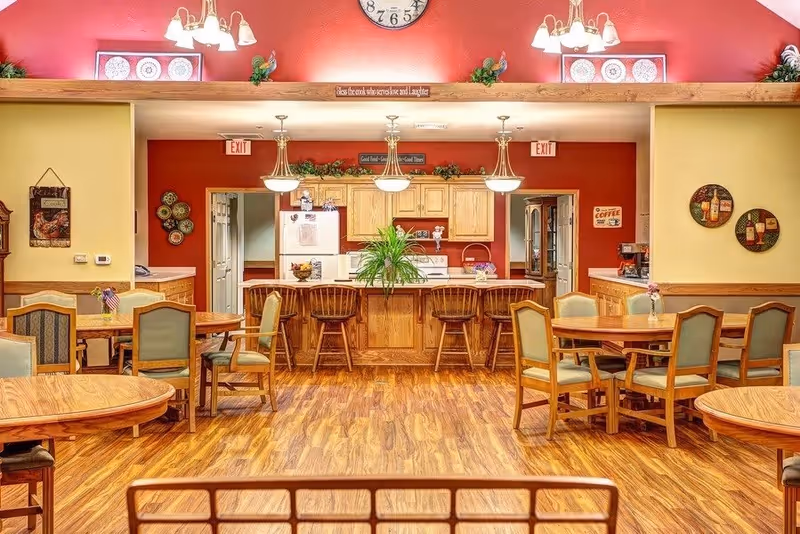 A warm and inviting communal dining area with wooden floors and multiple round wooden tables surrounded by green cushioned chairs. In the background, there is a kitchen area with wooden cabinets, a white refrigerator, a microwave, and a counter with four wooden bar stools. The walls are painted red and yellow, decorated with plates, signs, and small plants. Overhead lights hang above the kitchen counter, and a large clock is mounted on the wall above the open space between the dining area and kitchen.